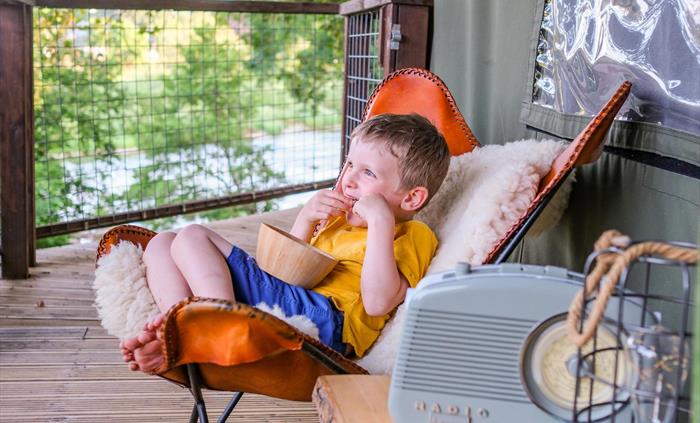 A child sat in a cozy chair on the decking of a safari tent eating popcorn watching the river.